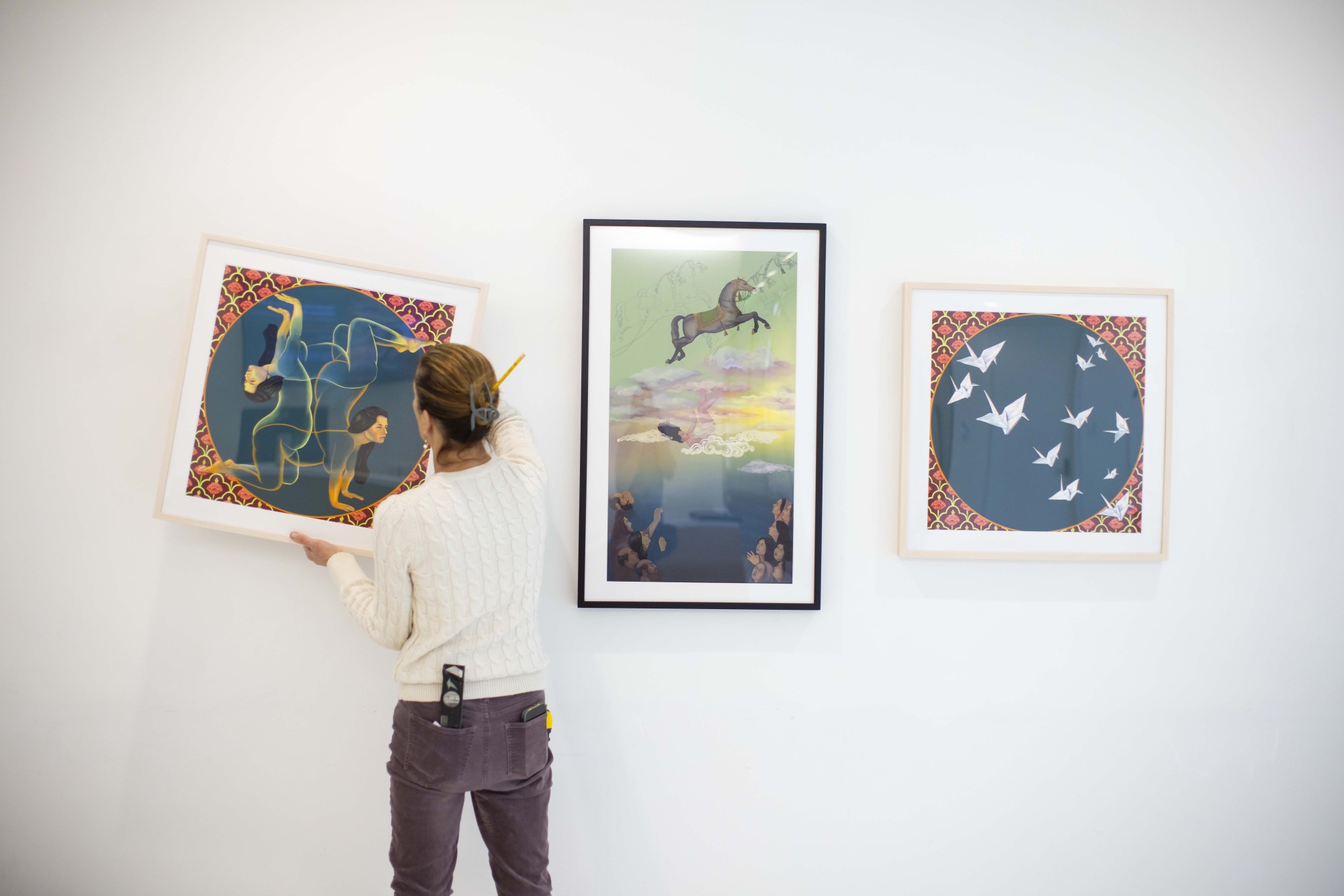 A person in a white sweater is adjusting a framed artwork on a gallery wall, flanked by two other vibrant, surreal artworks featuring intertwined figures, a floating horse, and origami cranes.