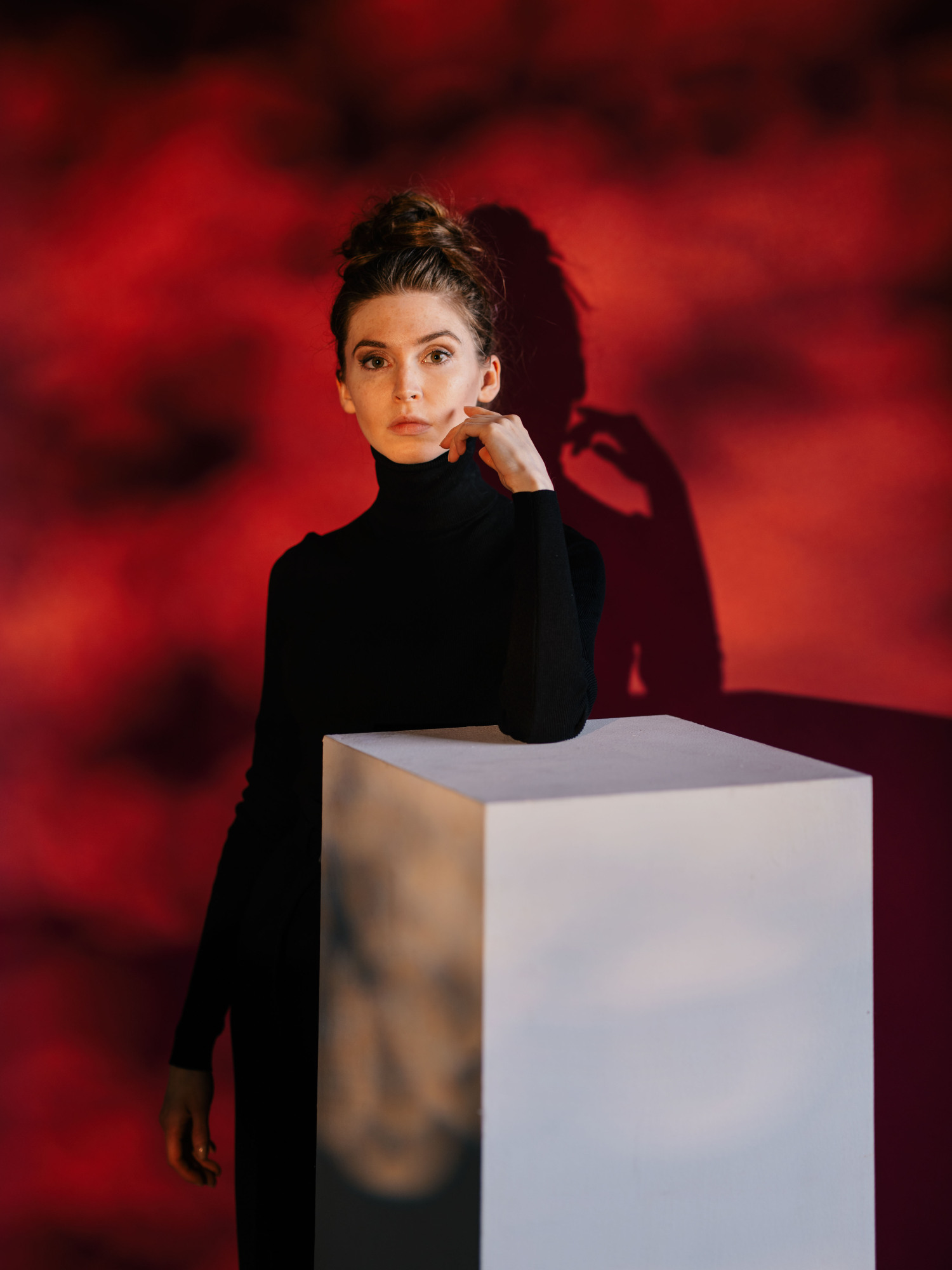A portrait of Jeanette Andrews in a black turtleneck leaning on a white pedestal against a textured red background.