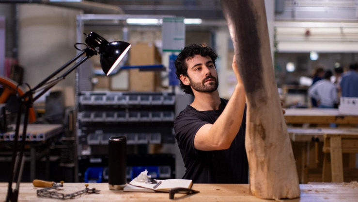A young man with curly dark hair and a beard named Eiden Spilker stands at a wooden workbench in a workshop carefully examining a large piece of raw timber while a bright desk lamp illuminates his tools and notebook