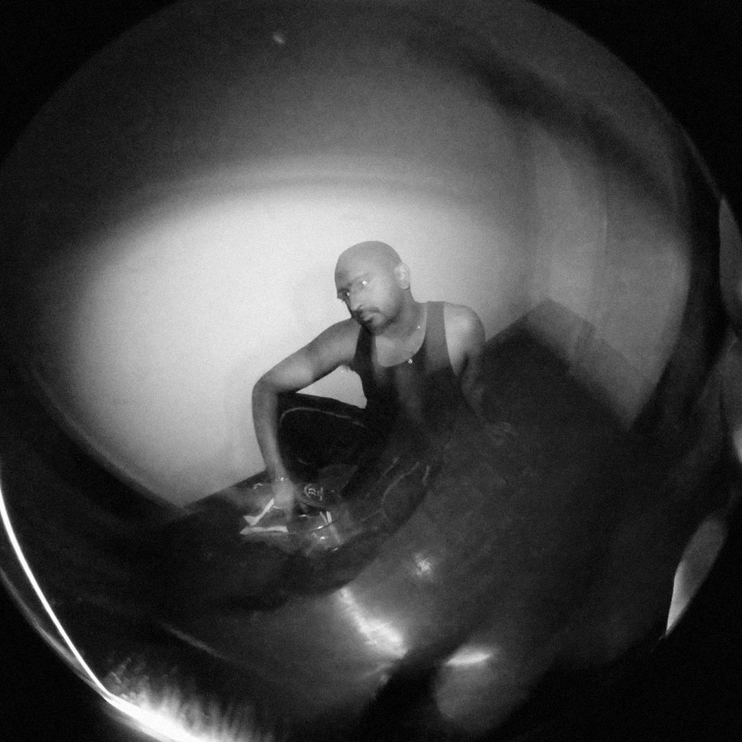 A black and white fisheye photograph of Rohan Chander crouching on the floor looking up with a hand on a drum