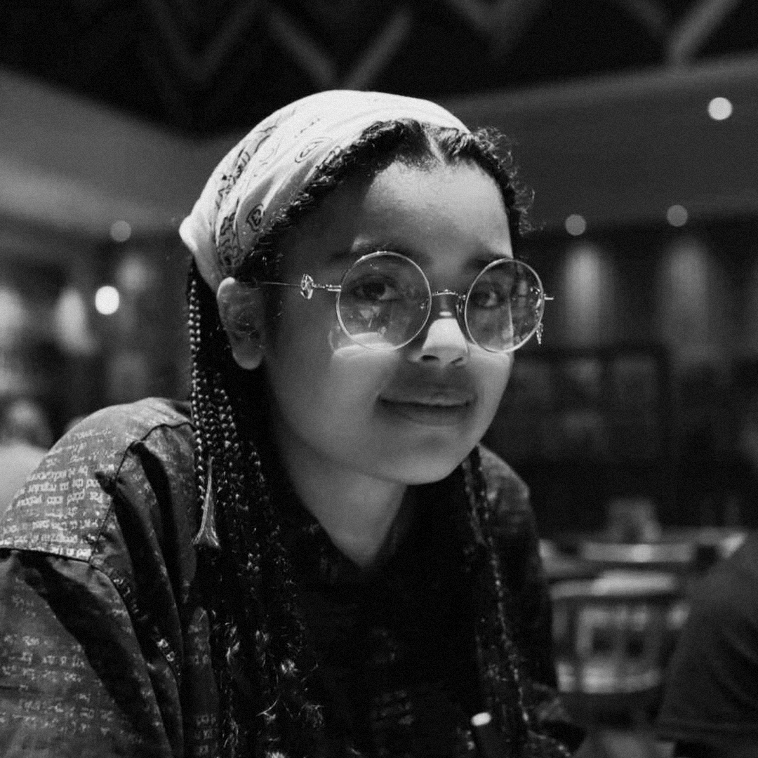 Cassius Hall with braided hair wearing round glasses and a patterned bandana smiles for a black and white portrait