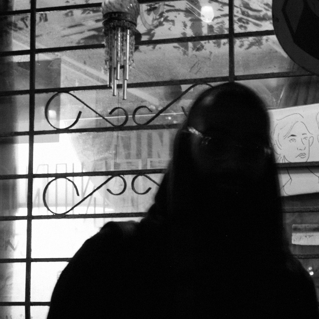 A deep silhouette of Vivek Menon stands before a gridded window with ornamental iron scrollwork and a hanging glass mobile and a sketched portrait is nearby