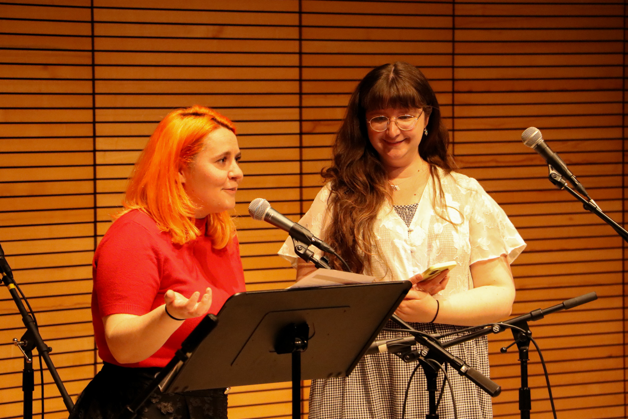 two people stand behind microphones in front of a horizontal wood slatted wall while the person with orange hair on the left gestures and speaks and the person on the right refers to a speech draft on their phone
