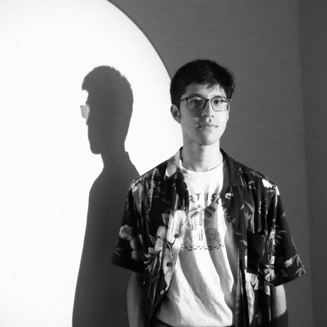 Luca Suarez wears a floral patterned shirt and glasses in a black and white portrait featuring a strong spotlight shadow on a wall