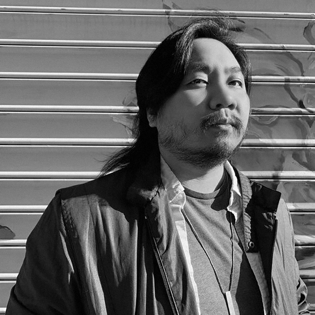 A black and white photograph of Phong Tran with long hair and a beard standing in front of a metal rolling door