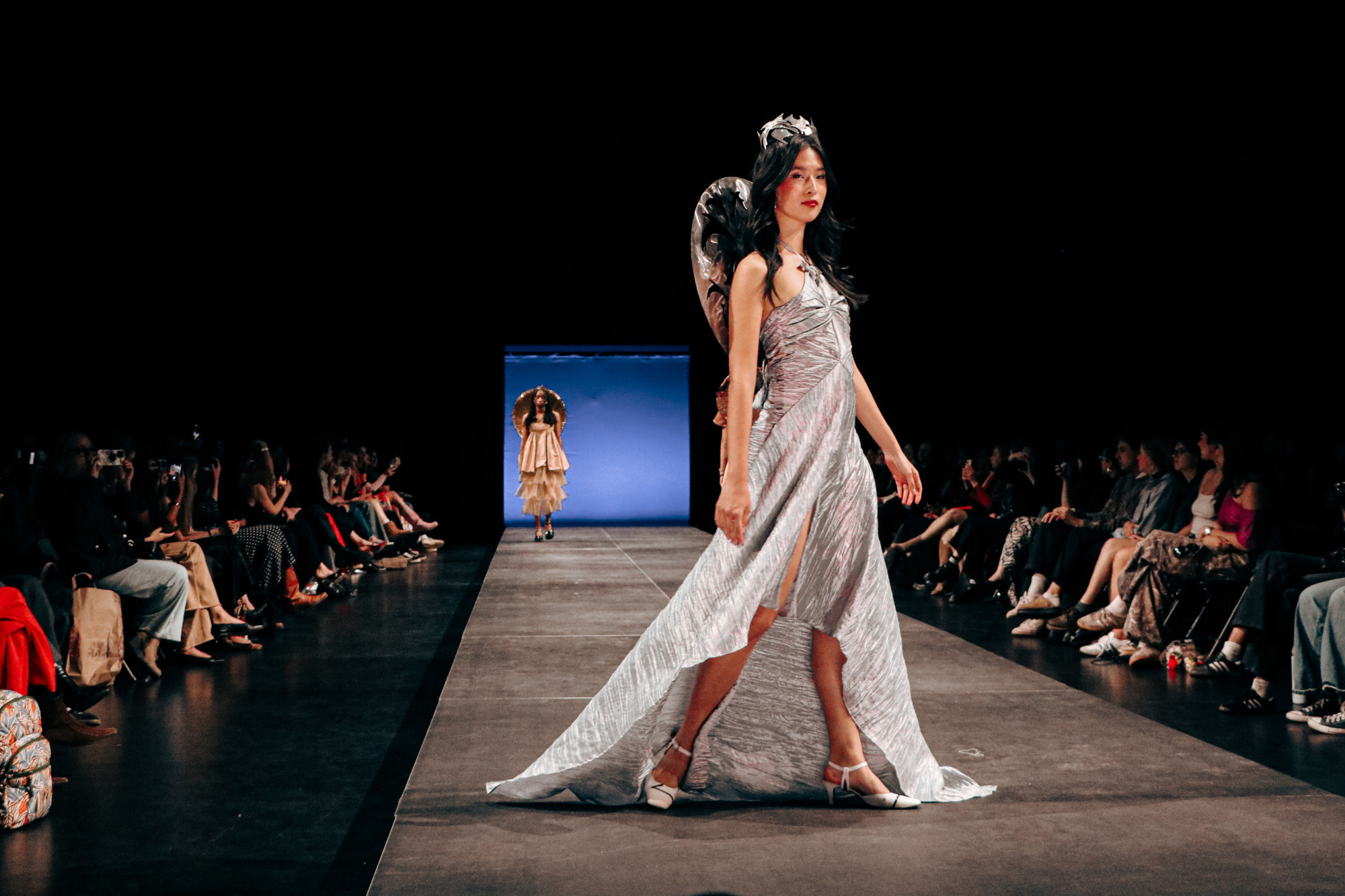 a model in a metallic silver dress and crown walks a runway during a fashion show while an audience watches from the sides