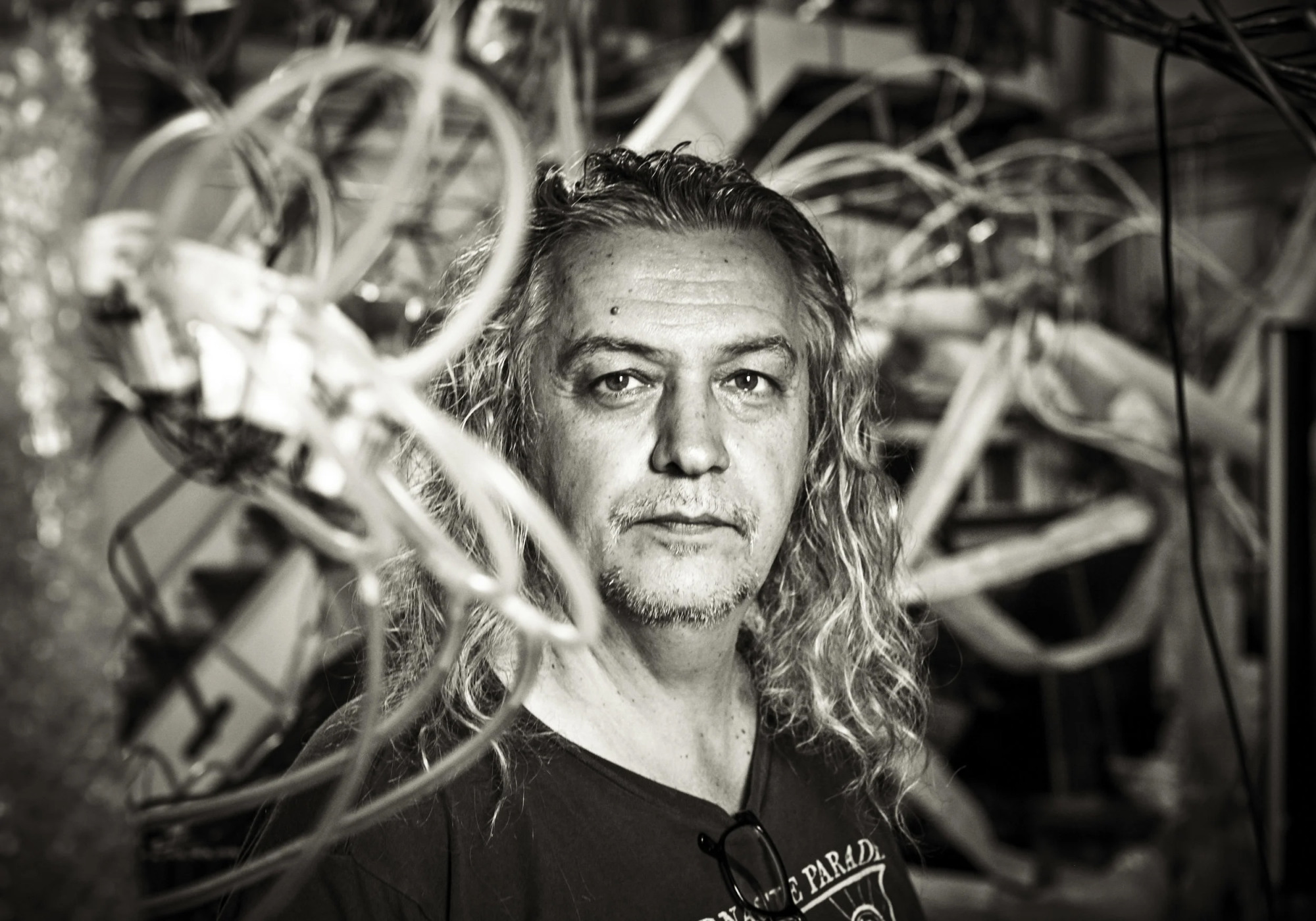A black-and-white portrait of a man with long, wavy hair looking directly into the camera while surrounded by complex, wire-like mechanical sculptures in a workshop setting.