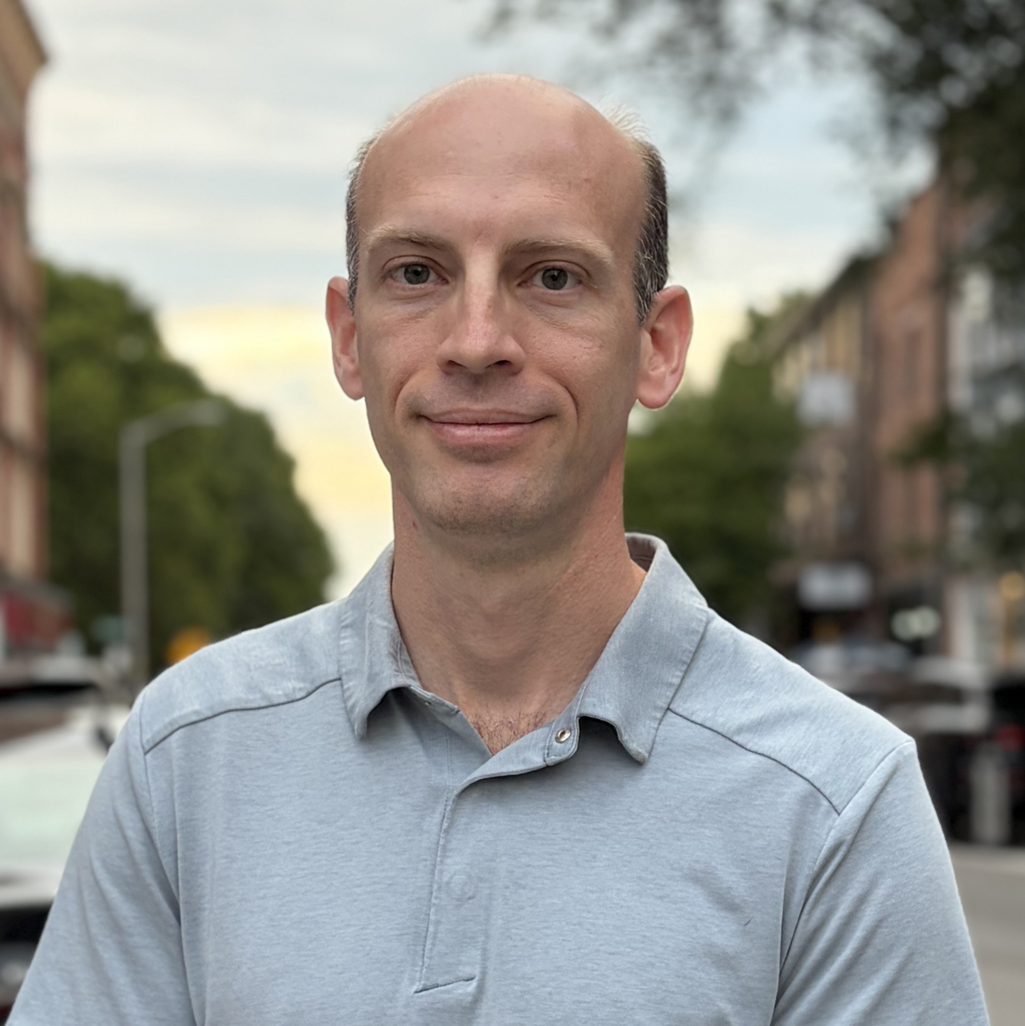 A headshot of Robert Goldschmidt smiling slightly outdoors, wearing a light gray collared polo shirt with a blurred street background.