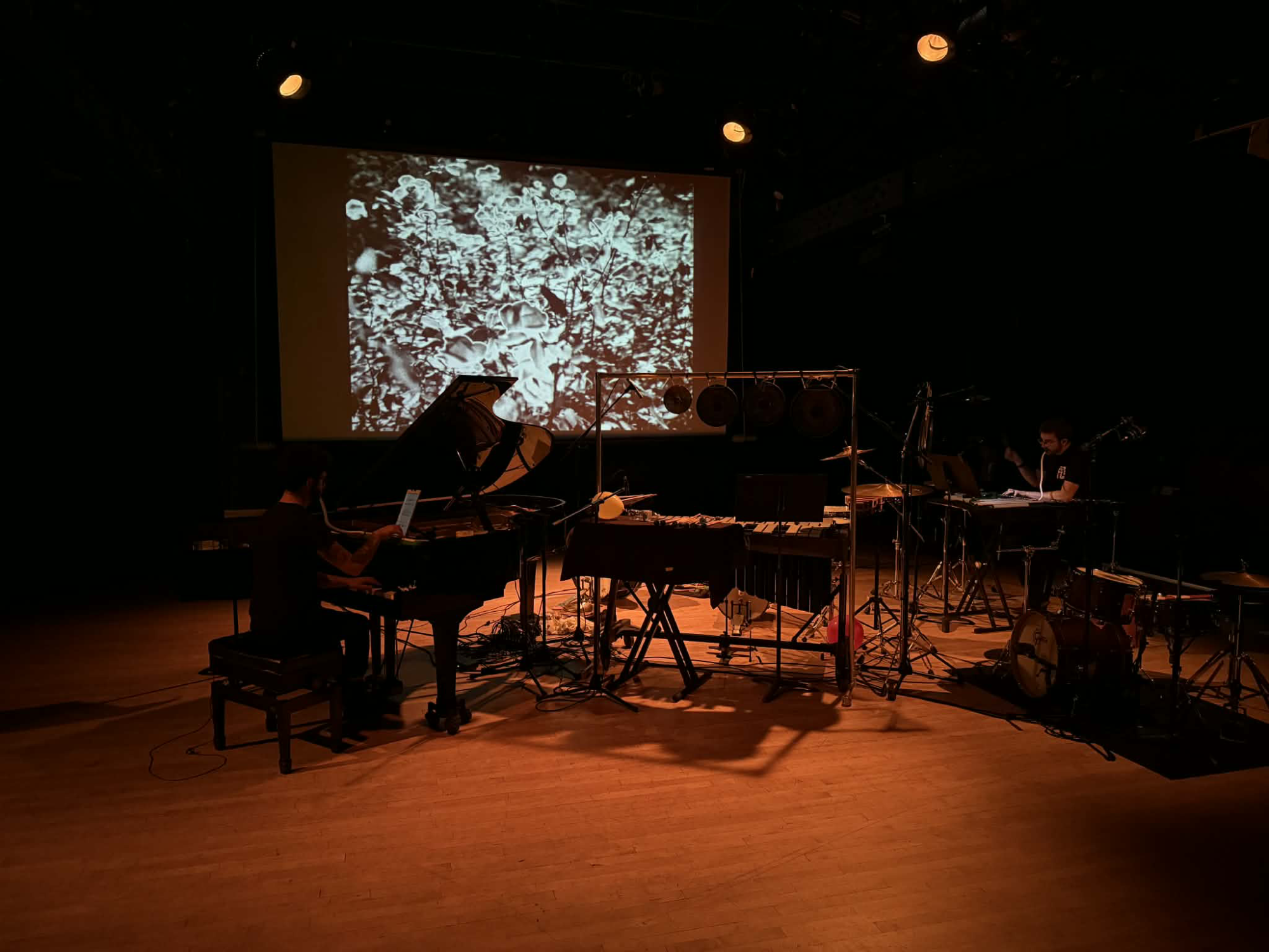 a dimly lit room with wood floors filled with instruments. two figures play piano and drums. a projection on a screen in the background shows an abstract texture in grayscale.