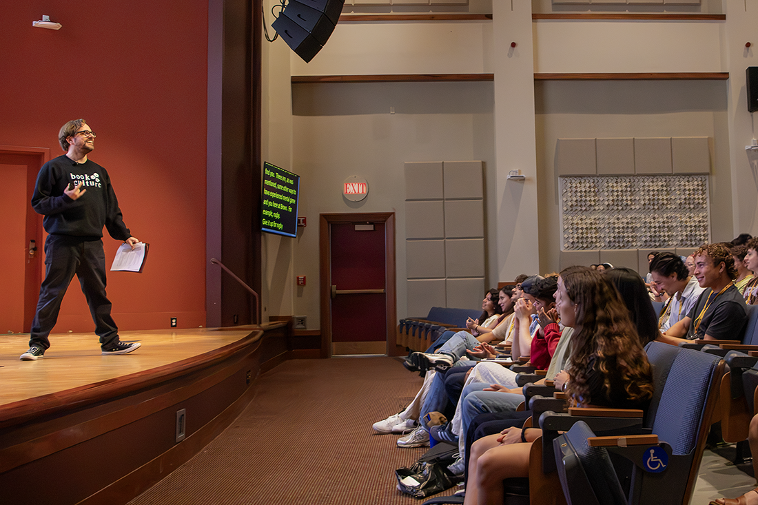 a smiling man with glasses stands onstage in front of an audience of students