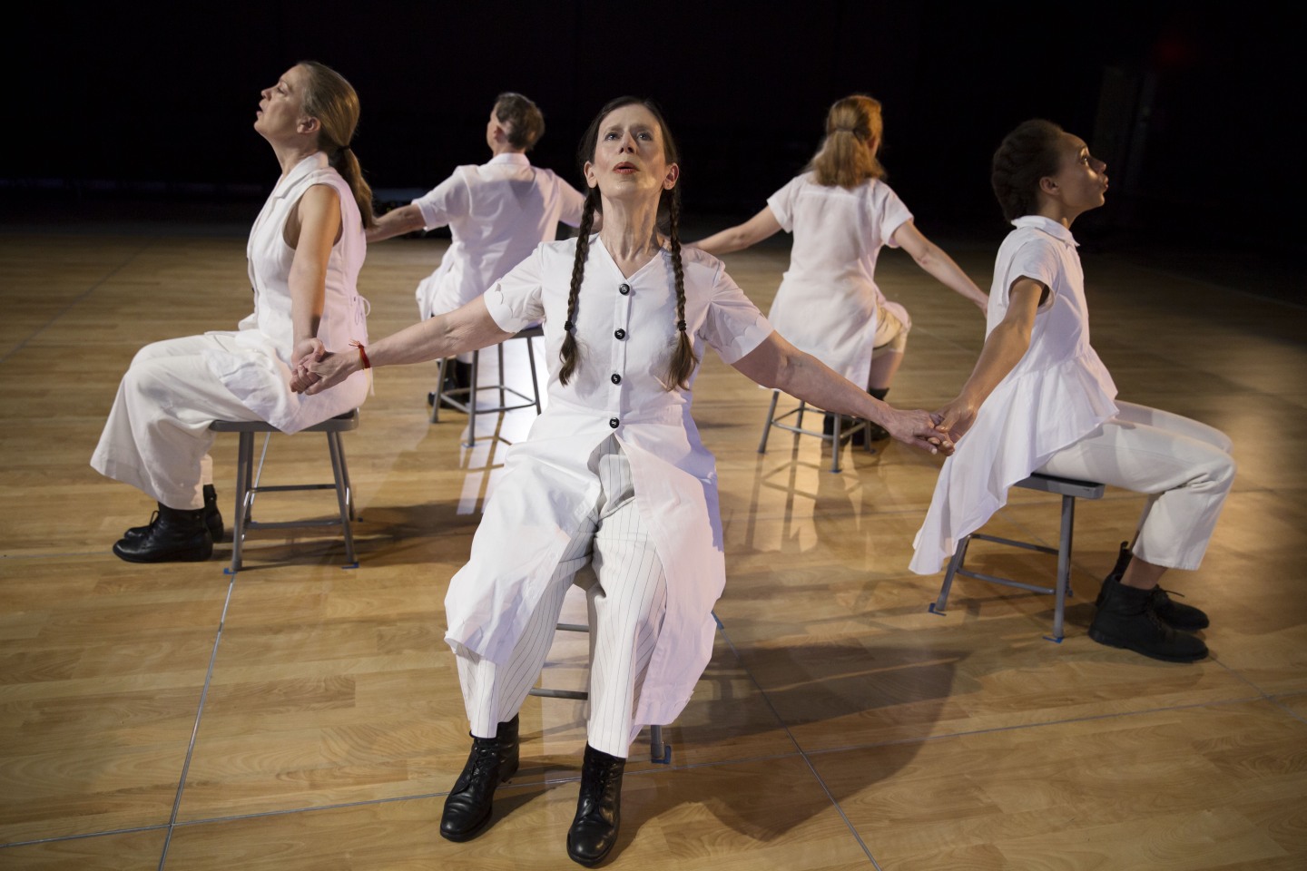 Meredith Monk and Vocal Ensemble sitting in chairs in a circle, facing out and holding hands.