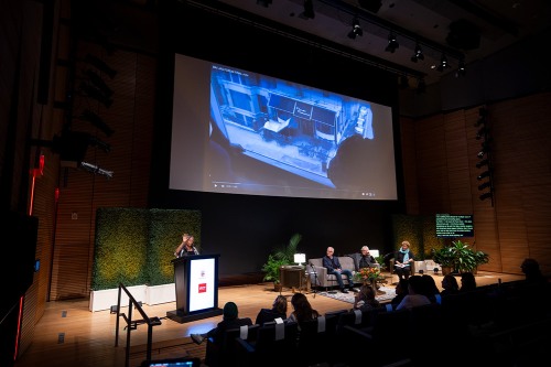A panel gives a presentation with a video screen in the background and a podium to the left