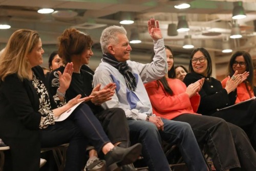 A number of people sitting in an audience clapping for a man who is raising his hand in recognition.