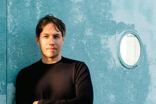 Sean Baker, a white man with dark hair, leans against a blue wall with a circular window.