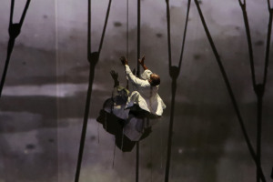 Performers in white garments on a dark stage with one performer suspended in mid-air against a large screen displaying abstract landscape projections and a web-like architectural shadow overlay