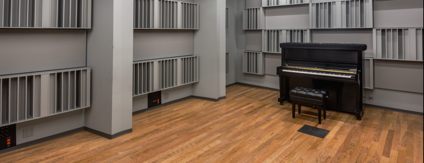 A view of the recording studio: wood floors, gray walls with audio dampening sections, an upright piano.