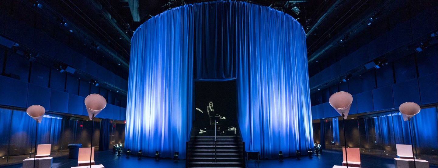 A large room with a circle of blue curtains in which a video image of woman in a sparkly aqua dress resides.