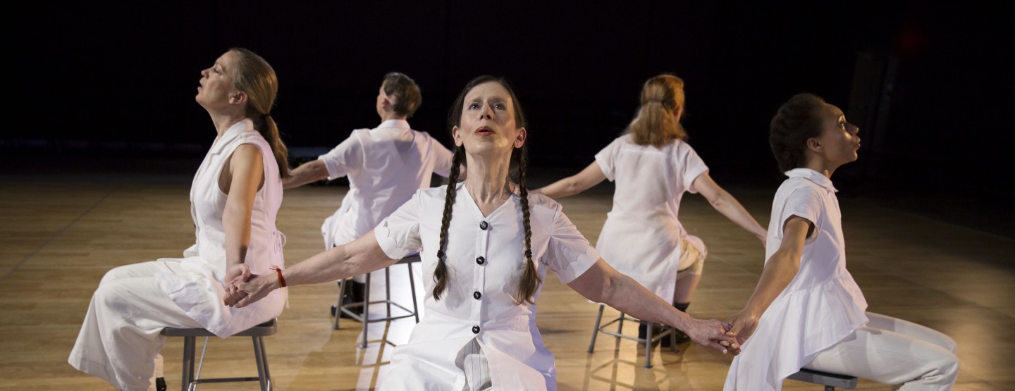 Meredith Monk and Vocal Ensemble sitting in chairs in a circle, facing out and holding hands.