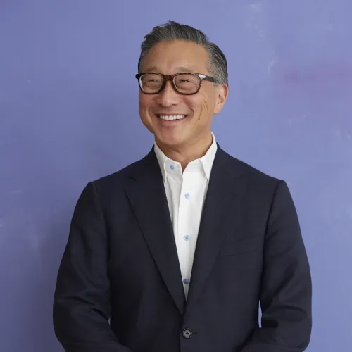 Emil Kang smiling in a dark suit with glasses on standing against a blue background