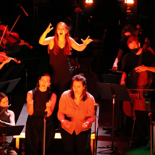 Three opera singers surrounded by an orchestra.