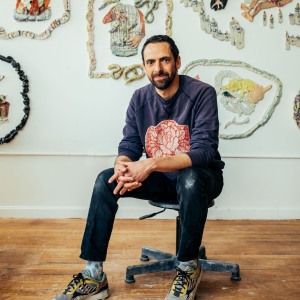 Judd Schiffman in a navy blue sweatshirt sitting on a rolling chair with his hands clasped looking at the camera with a serious expression