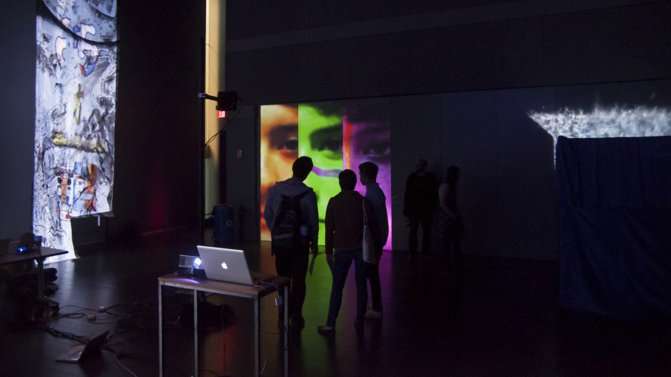 An installation with projections of a face on a wall and people in silhouette looking at it. A computer sits on a table in the foreground.