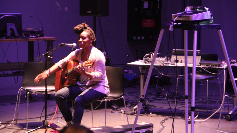 A musician performing with a guitar sitting in front of microphone. A projector is in the foreground.