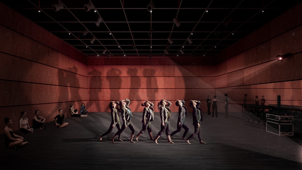 A group of dancers move in a line with their hands to their heads. A beam of light from the right sends their shadows to the back wall.