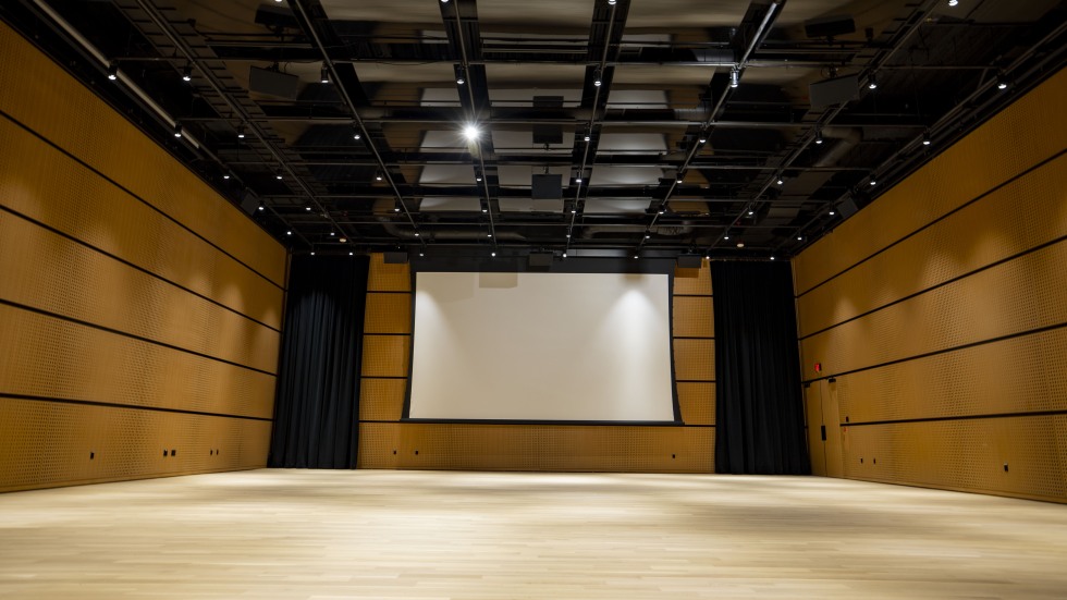 An empty rehearsal hall with wood flooring and walls and a large projection screen.