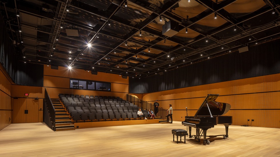 A grand piano on a stage floor with seating in the background. Four people in conversation.