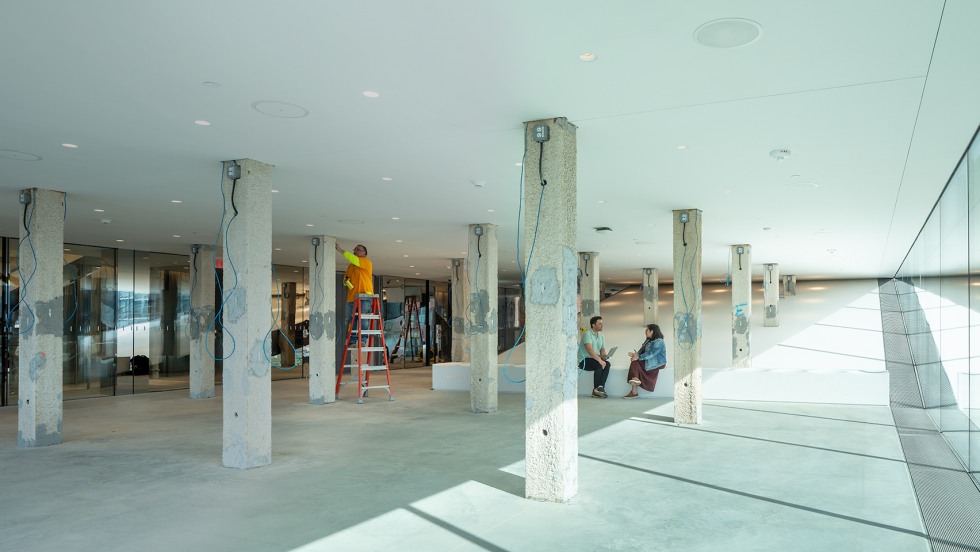 Two people seated on a bench are engaged in conversation. The room has white walls, ceilings, and pillars.