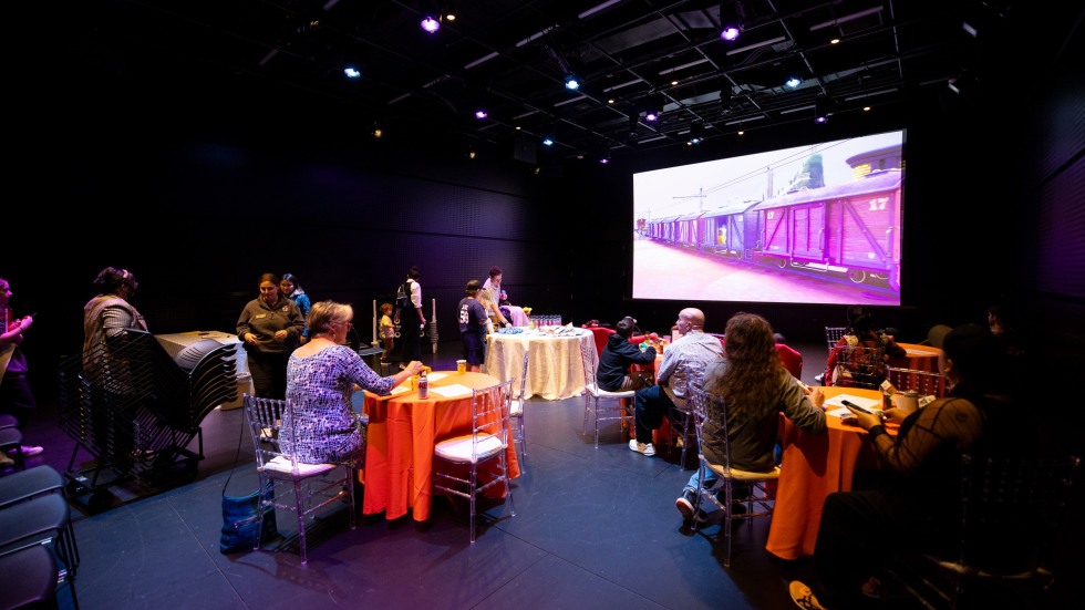 dimly lit room with round tables and chairs; parents and children watching a movie on a projections screen