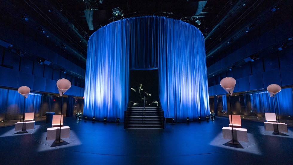 A large room with a circle of blue curtains in which a video image of woman in a sparkly aqua dress resides.