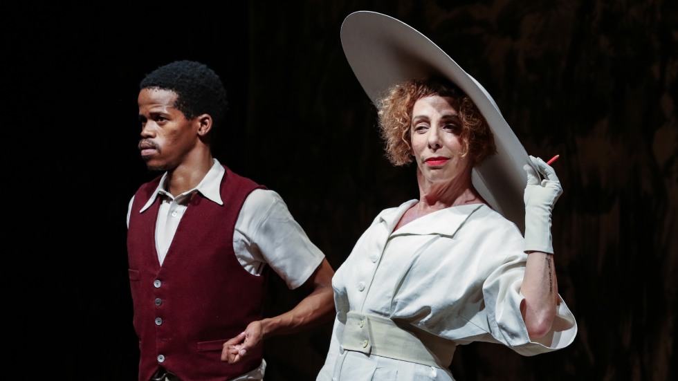 An image from Houseboy. A young man in a red vest stands next to a woman in a white dress and large hat