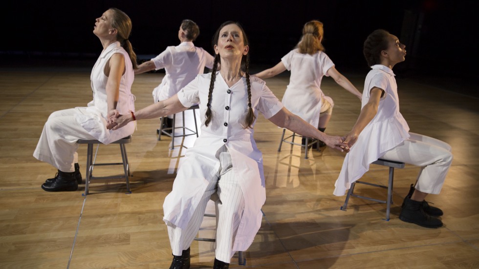 Meredith Monk and Vocal Ensemble sitting in chairs in a circle, facing out and holding hands.