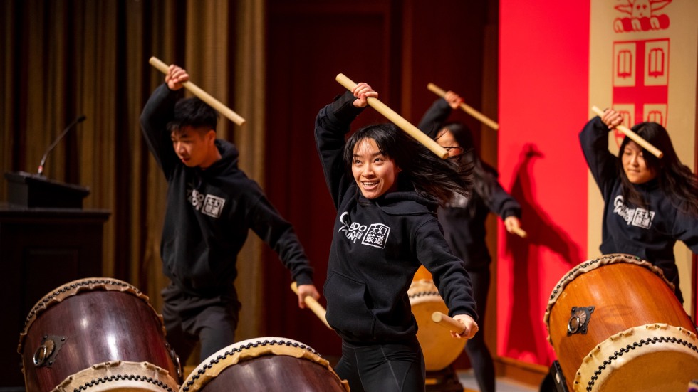 Taiko drummers perform. All four have drumsticks raised above their heads.