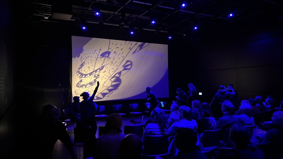 People seated in chairs watching a performance with a drawing projected on a large screen.