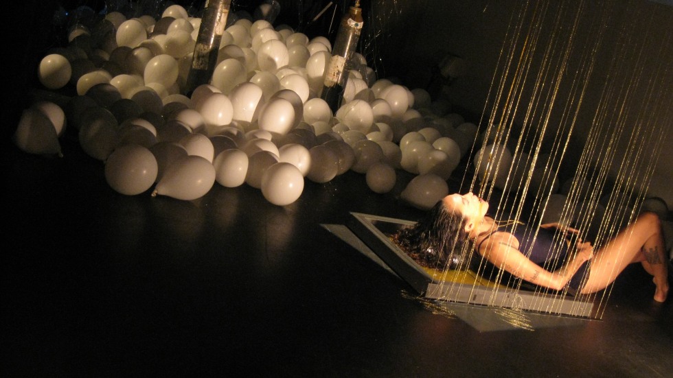 Julie Tolentino performing Honey on a dark floor with white balloons to her left