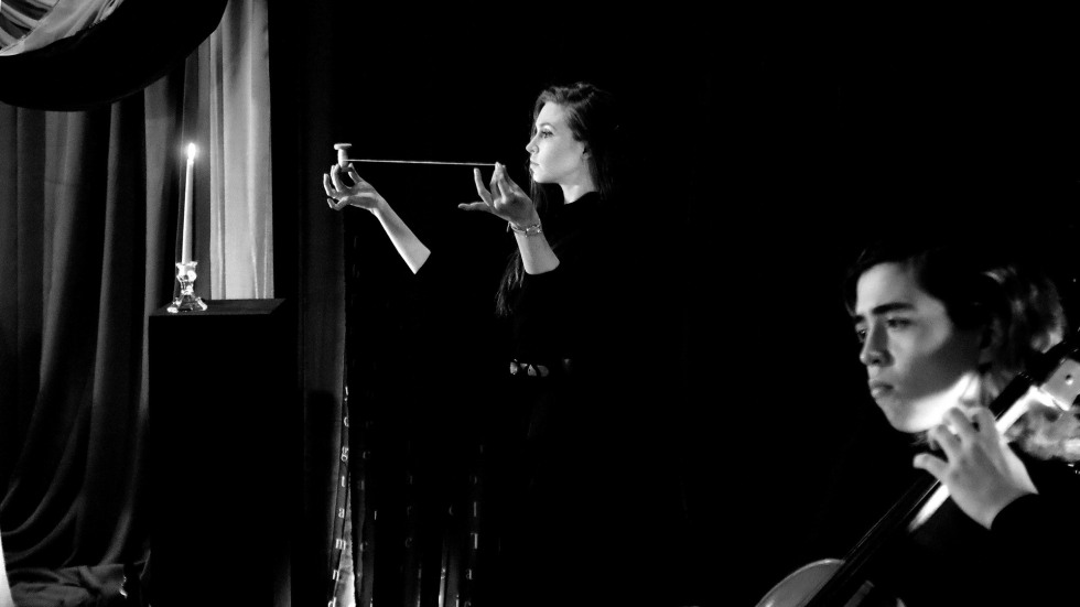 A black and white photograph of a theatrical performance. In the background, a woman in a dark dress holds a horizontal string or wire between her hands with focused intensity. To her left, a single lit candle sits on a tall black pedestal. In the blurred foreground, a cellist is seen from the side, mid-performance, creating a sense of depth and atmosphere.