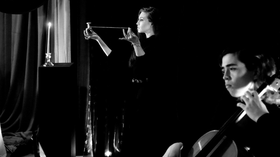 A black and white photograph of a theatrical performance. In the background, a woman in a dark dress holds a horizontal string or wire between her hands with focused intensity. To her left, a single lit candle sits on a tall black pedestal. In the blurred foreground, a cellist is seen from the side, mid-performance, creating a sense of depth and atmosphere.