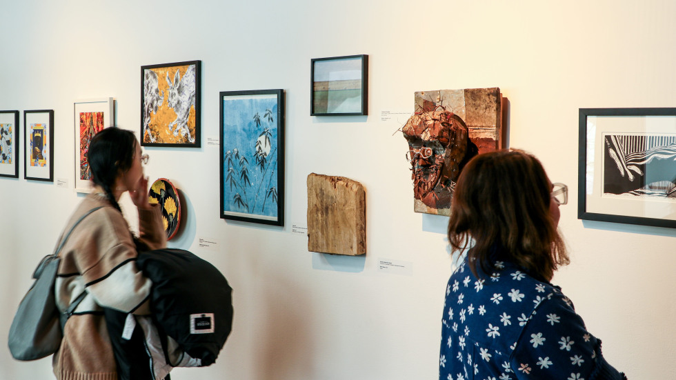 An indoor gallery scene showing a white wall filled with various art pieces at different heights. To the left, a person with a backpack looks at a framed yellow and white illustration of animals. The center of the wall features a blue bamboo painting and a 3D textured mask of a face. To the right, another person in a blue floral shirt observes a black-and-white abstract print. The lighting is soft, highlighting the textures of the individual artworks.