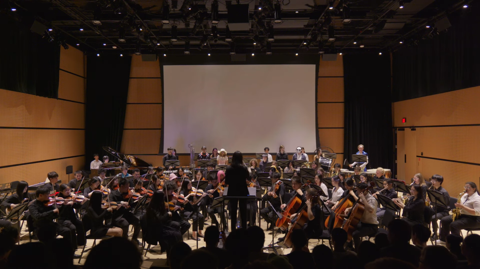 A medium-shot photograph of a diverse orchestra on a stage with wood-paneled walls playing instruments like violins and cellos under warm lights and with a blank projection screen in the background.