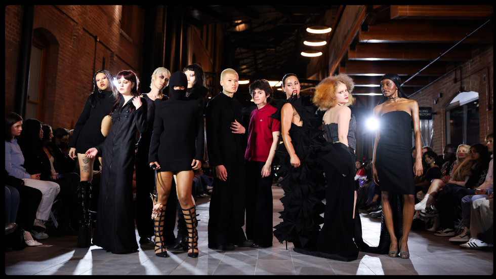 A group of fashion models wearing avant-garde black garments and one red polo shirt posing on a runway in an industrial brick building