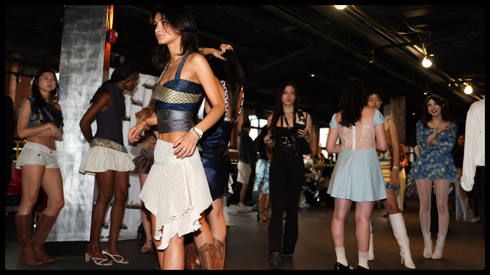 Multiple fashion models preparing backstage at a runway event wearing diverse outfits including a top made of neckties and a white asymmetrical skirt