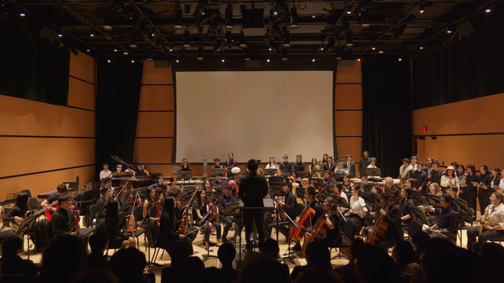 A medium-shot photograph of a diverse orchestra on a stage with wood-paneled walls playing instruments like violins and cellos under warm lights and with a blank projection screen in the background.