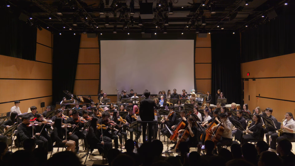 A medium-shot photograph of a diverse orchestra on a stage with wood-paneled walls playing instruments like violins and cellos under warm lights and with a blank projection screen in the background.