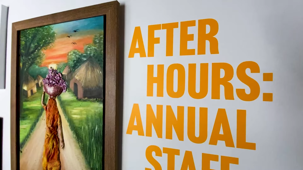 exhibition wall featuring large orange text for after hours annual staff next to a framed painting of a person walking through a village under a sunset sky