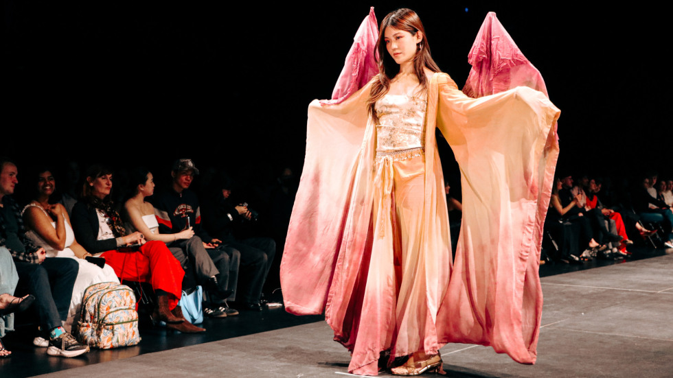 a model in a flowing yellow and pink gradient outfit with oversized sleeves poses on a runway during a fashion show