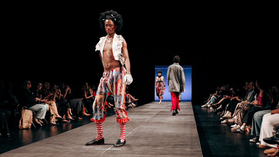 a model with an afro in patterned pants and striped socks poses on a runway while an audience watches from the sides