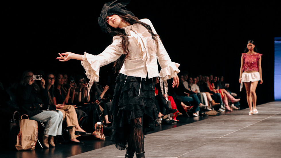 a model in a ruffled white blouse and tiered black skirt poses on a runway while an audience watches
